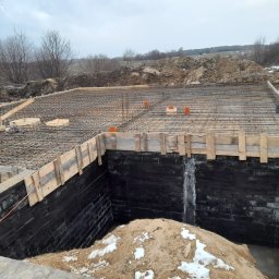 Tombud - Zabezpieczone, czarne ściany fundamentowe z drewnianym szalunkiem i zbrojeniem na górze. W tle ziemia i pochmurne niebo. Widoczne elementy instalacji.