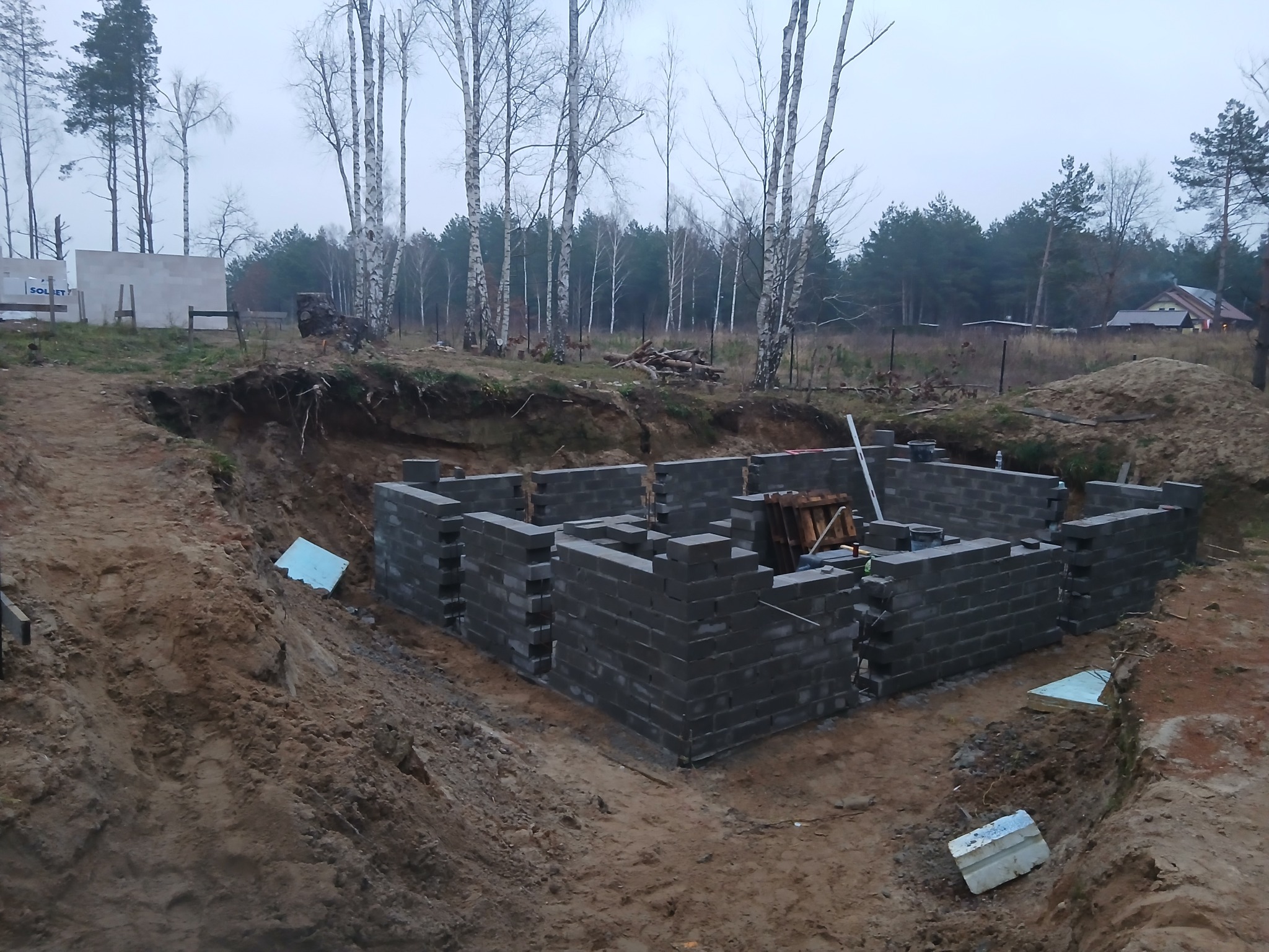 Wykop pod fundamenty z częściowo wymurowanymi ścianami z szarego bloczka betonowego. W tle drzewa i budynek w oddali. Widoczne narzędzia i materiały budowlane.