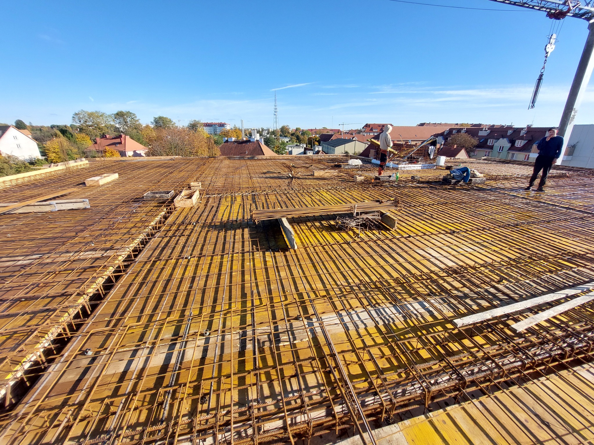 Zbrojenie stropu budynku w trakcie budowy, widoczne żółte szalunki i stalowe pręty. W tle zabudowa miejska i błękitne niebo.