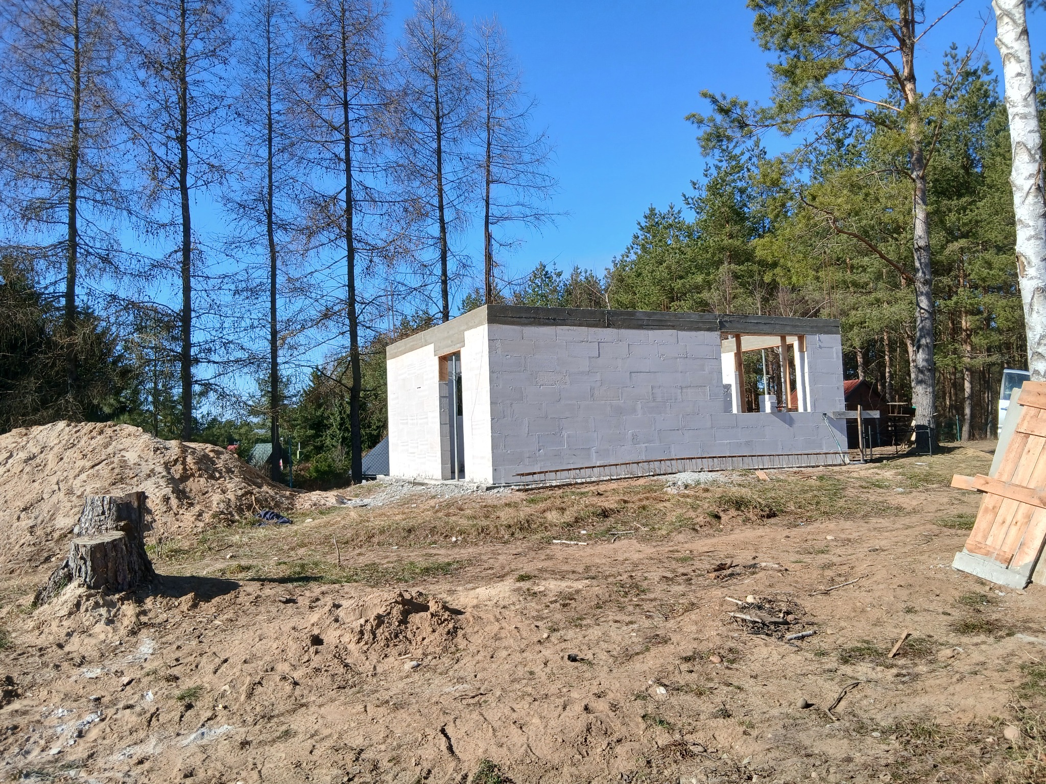 Wznoszenie ścian parterowego domu z betonu komórkowego na fundamencie, widoczne zbrojenie i otoczenie budowy z drzewami w tle.