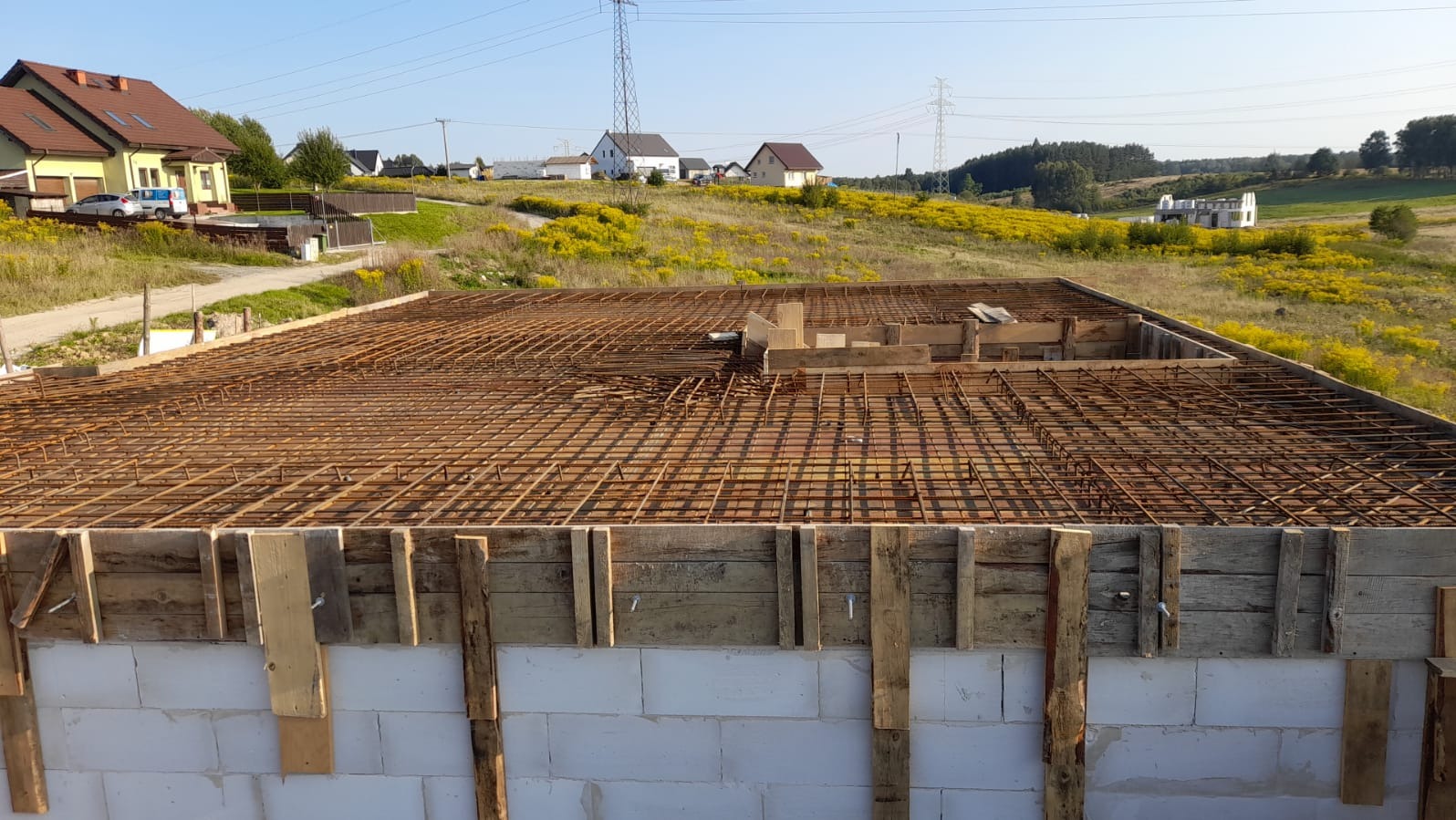 Zbrojenie płyty fundamentowej na budowie domu jednorodzinnego. Widoczne białe bloczki fundamentowe, drewniane szalunki i siatka zbrojeniowa. W tle zabudowa wiejska.