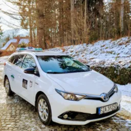 Biała taksówka kombi Toyota Auris Hybrid na brukowanej drodze w zimowej scenerii. Widoczny numer rejestracyjny FZA 59250 i oznaczenie TAXI.