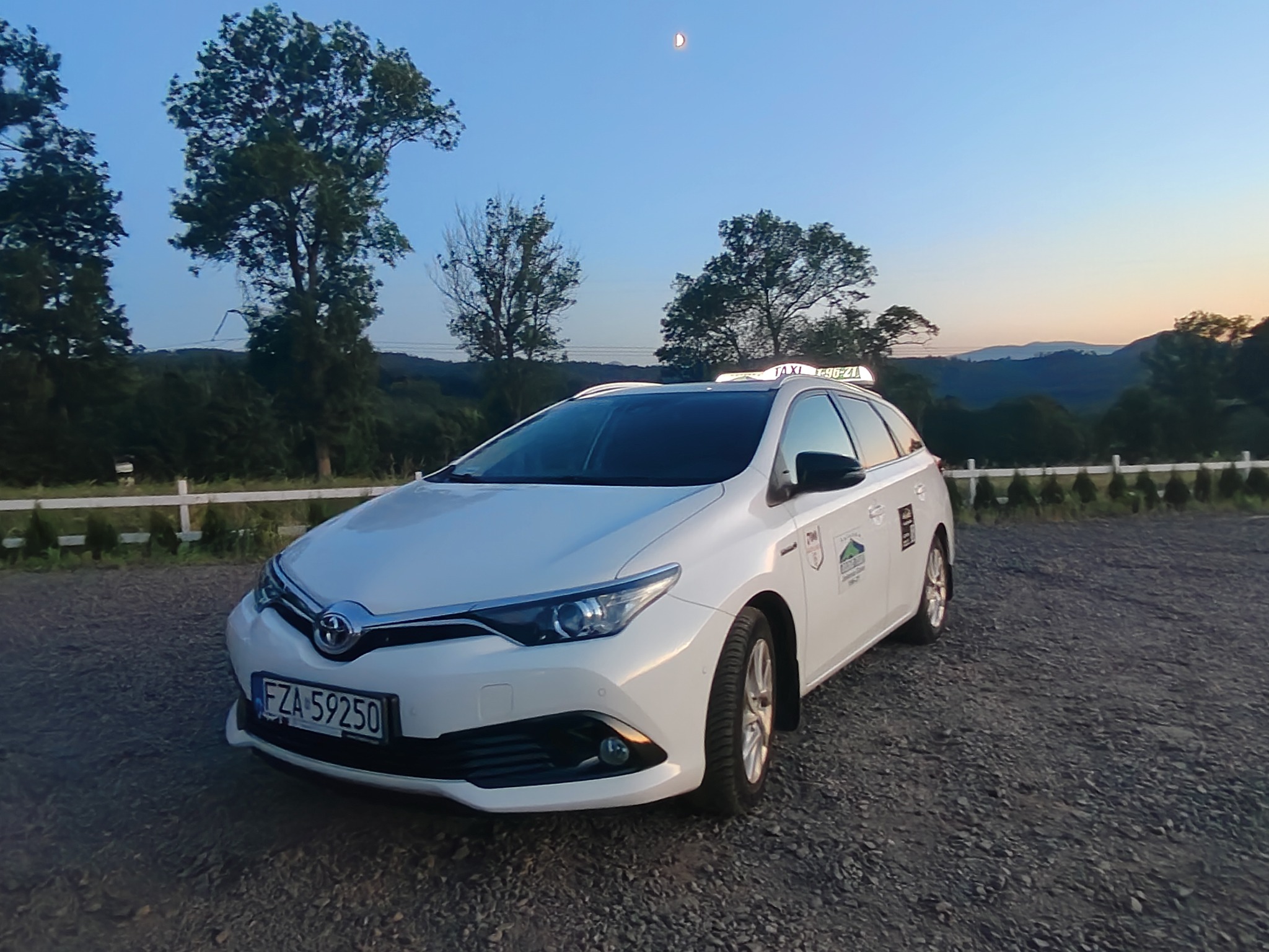 Białe kombi taxi na tle gór i zachodzącego słońca. Na dachu neonowy napis 'TAXI'. Widoczny numer rejestracyjny FZA 59250.