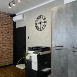 Nowoczesne biuro: drukarka Develop ineo 227, zegar na ścianie, ceglana ściana, szafki w odcieniach szarości, oświetlenie sufitowe i klimatyzacja.