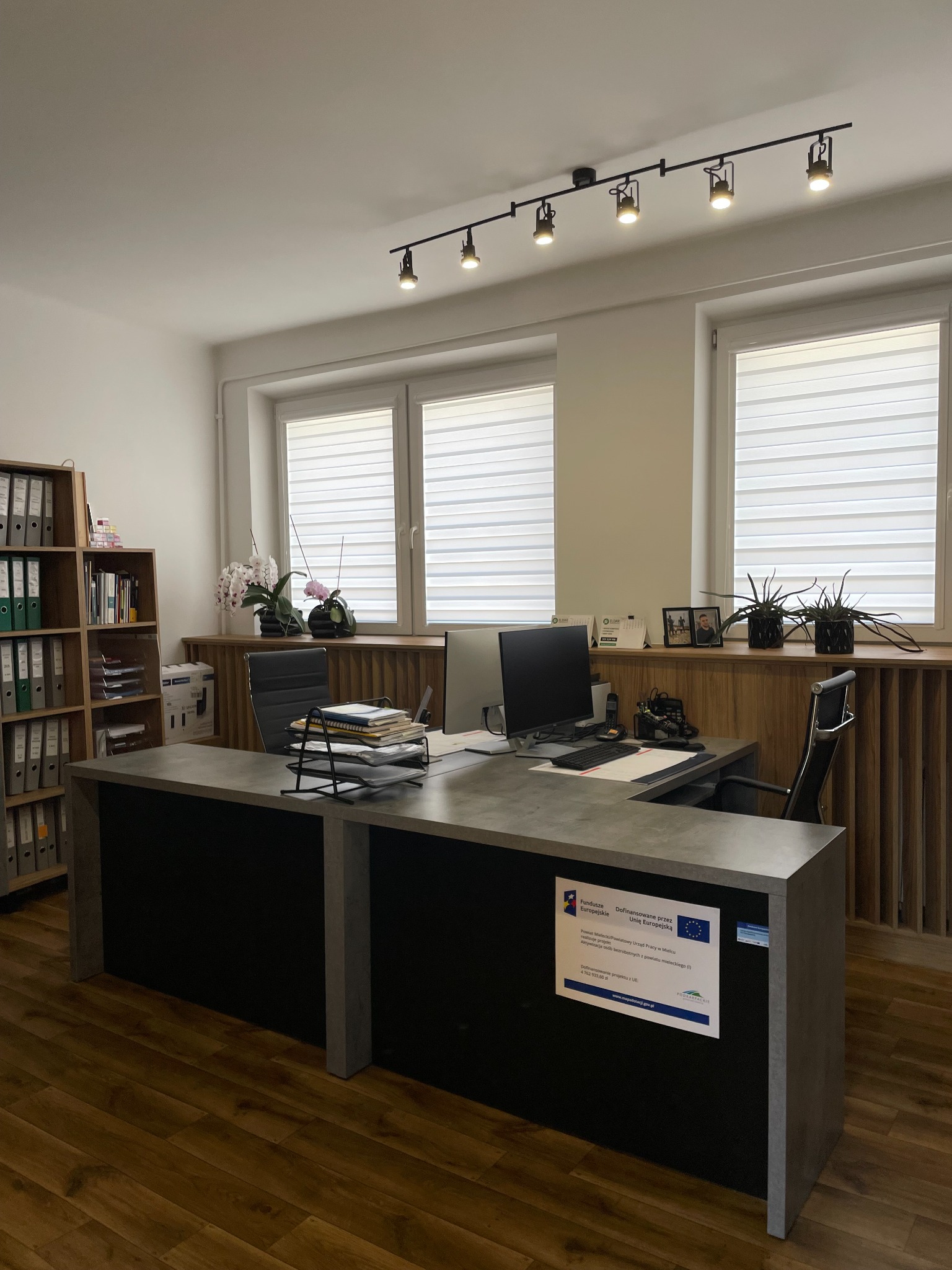 Jasne biuro z biurkiem w kolorze betonu, regałem na dokumenty i oknami z żaluzjami. Na biurku komputer, lampka i dokumenty. Na ścianie plakat z informacją o dofinansowaniu UE.