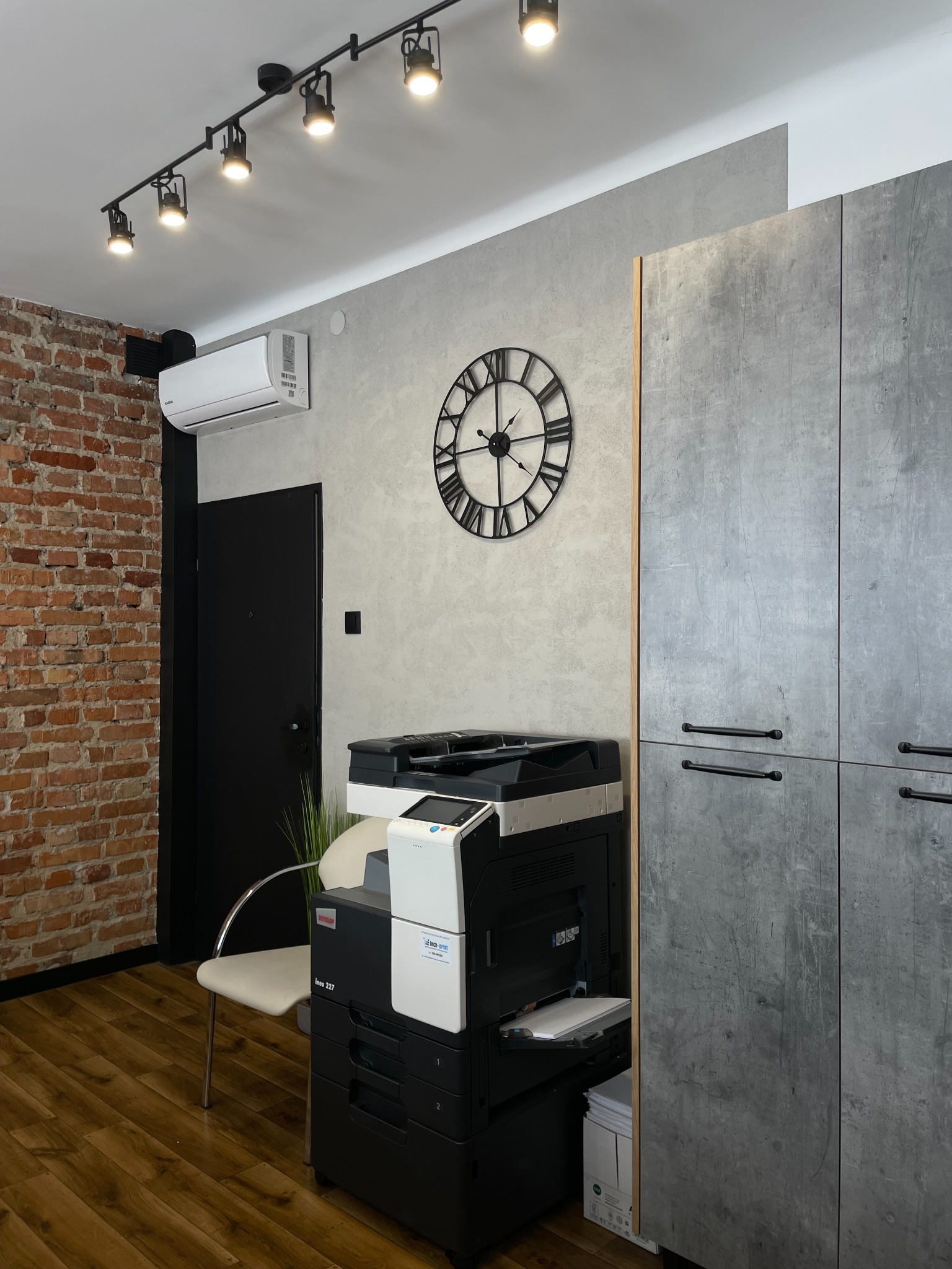 Nowoczesne biuro: drukarka Develop ineo 227, zegar na ścianie, ceglana ściana, szafki w odcieniach szarości, oświetlenie sufitowe i klimatyzacja.