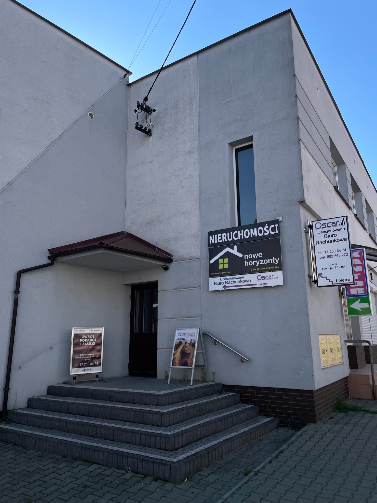 Budynek biurowy z szyldami 'Nieruchomości nowe horyzonty' i 'Oscar. Licencjonowane Biuro Rachunkowe' oraz tablicą informującą o zwrocie podatku.