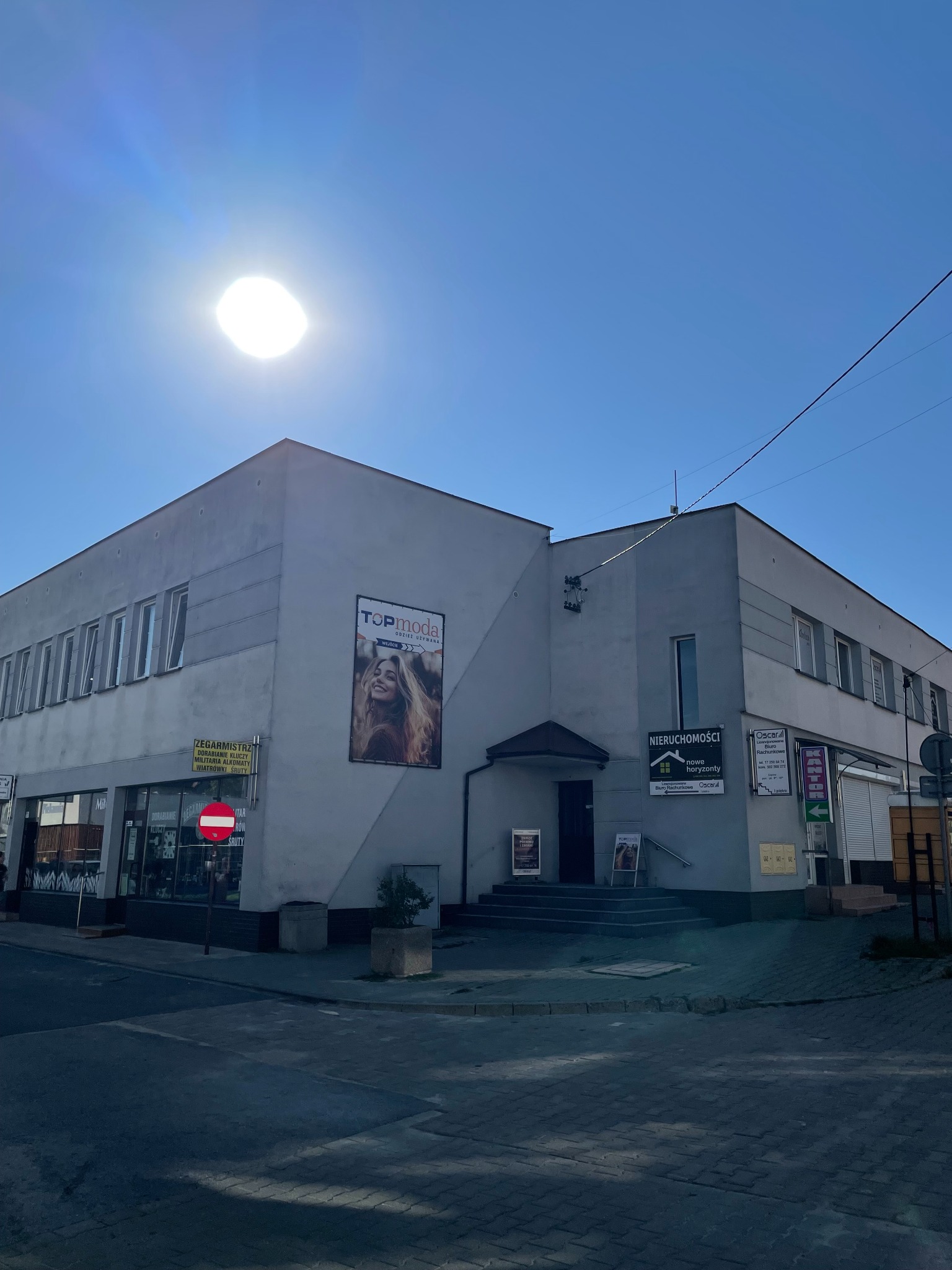Dwupiętrowy budynek biurowy z szyldami firm (nieruchomości, biuro rachunkowe, odzież używana) w słoneczny dzień. Widoczne reklamy na fasadzie.