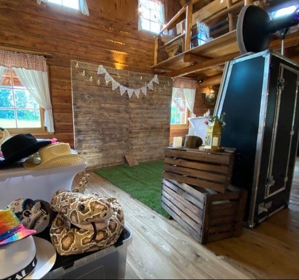 Rustykalna scenografia z drewnianą ścianką, girlandą i rekwizytami (kapelusze, pluszowy wąż) obok fotobudki w drewnianym wnętrzu.