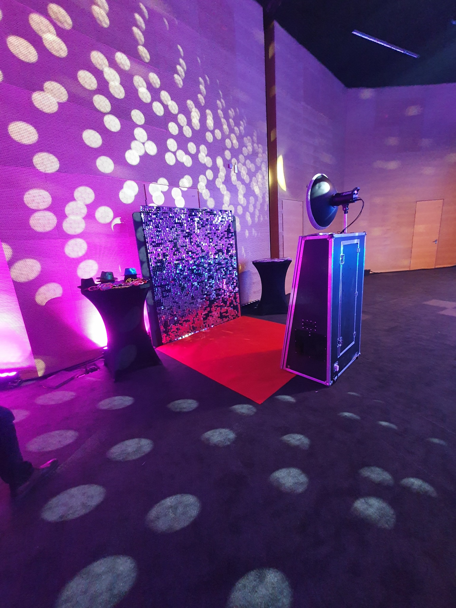Instalacja fotobudki z błyszczącym tłem, rekwizytami na stoliku i reflektorem, oświetlona fioletowym światłem. Czerwony dywan dopełnia całość.
