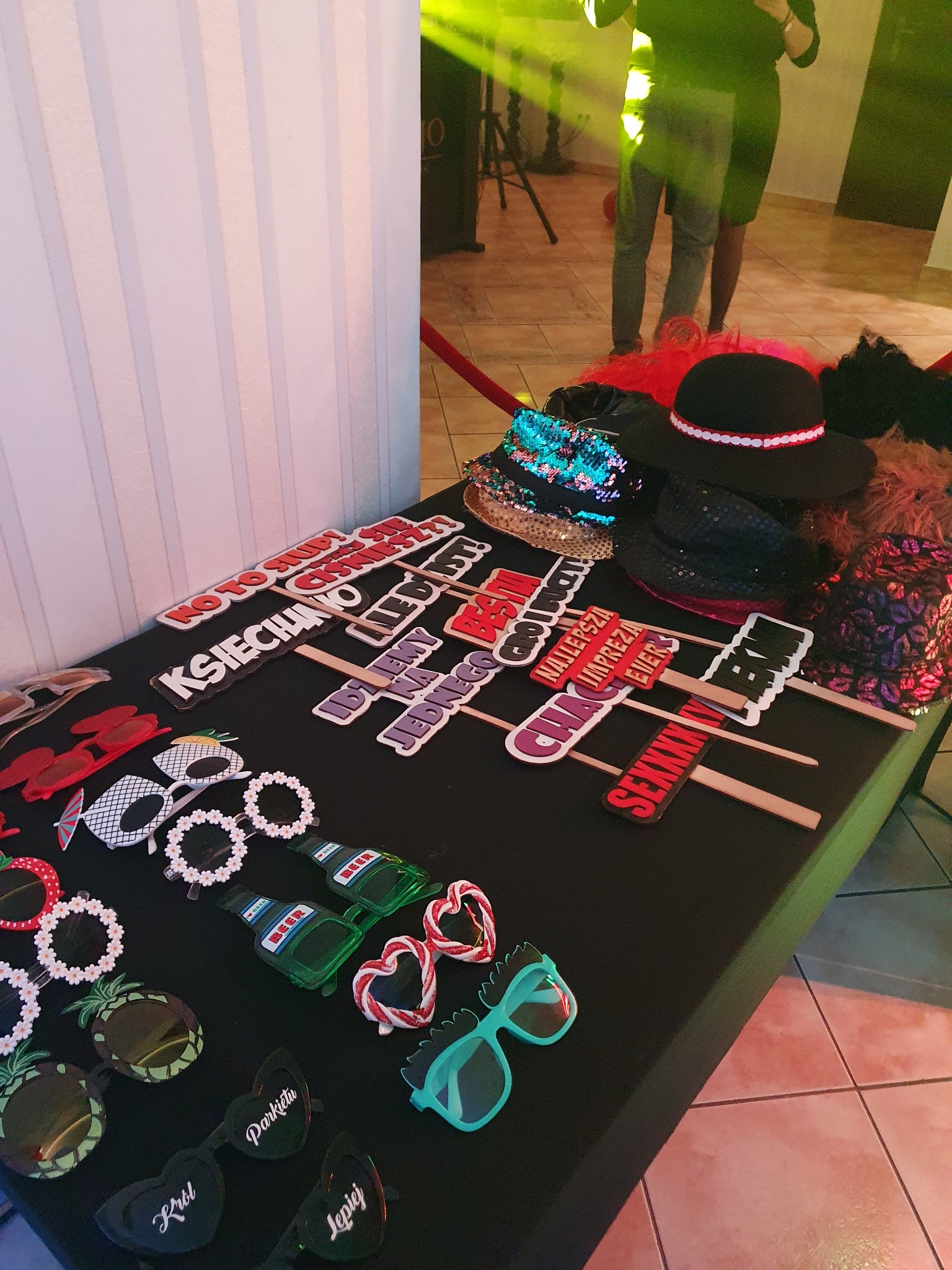 Akcesoria do fotobudki: okulary, kapelusze i tabliczki z napisami, rozłożone na stole z czarnym obrusem. W tle osoby i oświetlenie imprezowe.
