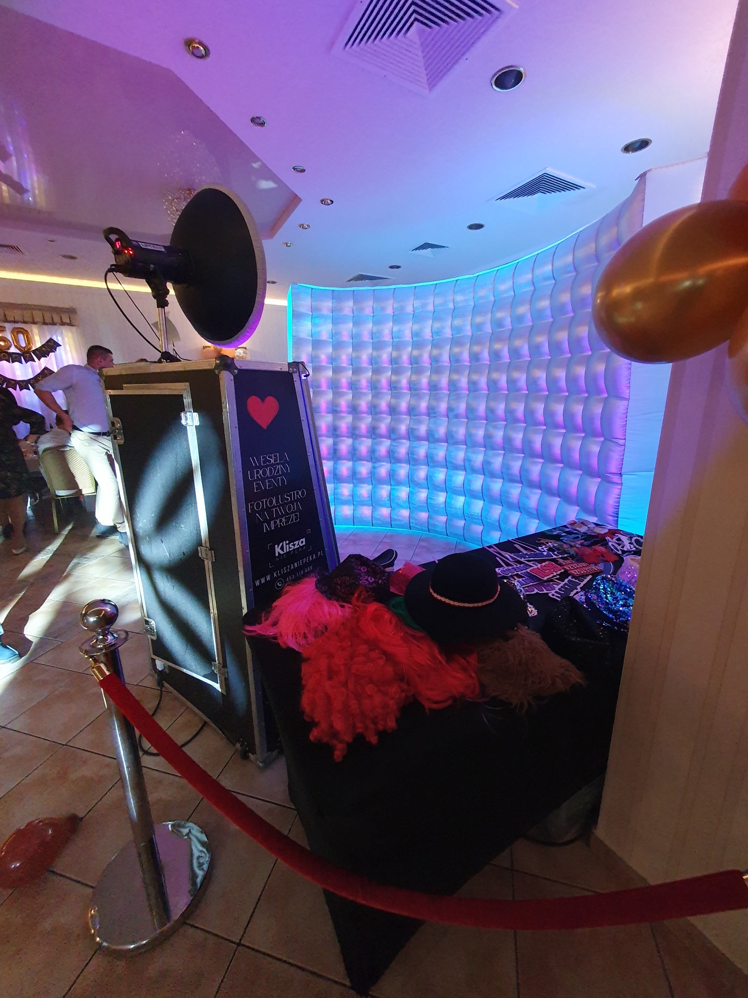Fotobudka z rekwizytami (peruki, kapelusze) i podświetlanym tłem na imprezie. Widoczny słupek z czerwonym sznurem wyznaczający strefę. Częściowo widoczni goście.