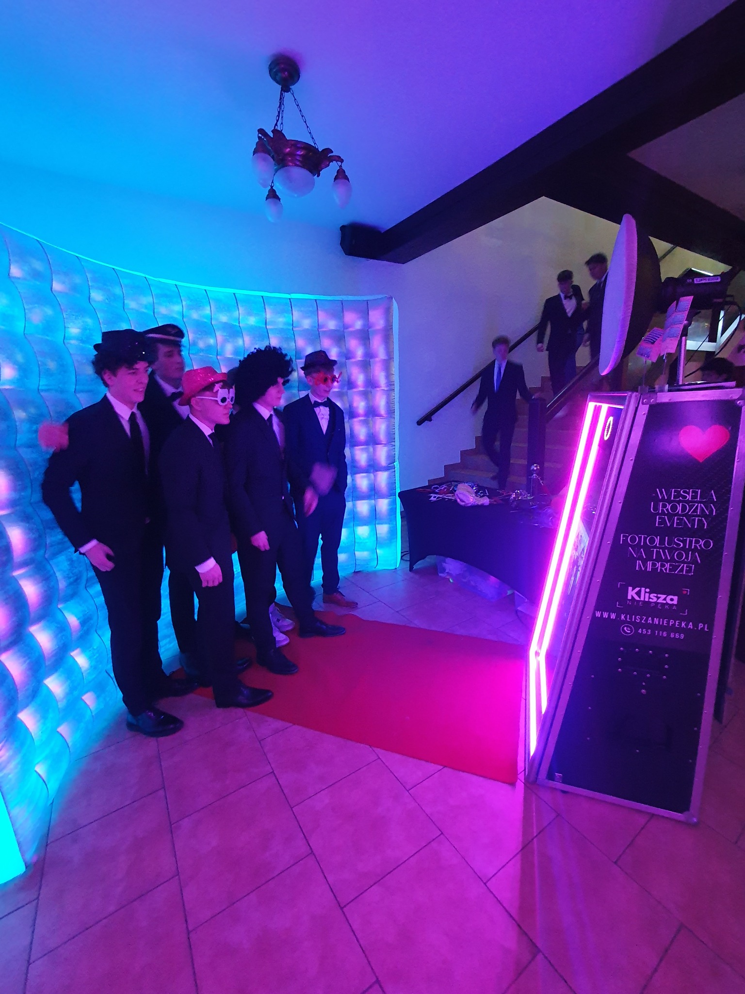 Grupa osób w przebraniach pozuje do zdjęcia w fotobudce z neonowym oświetleniem, dekoracyjna ściana w tle. Na schodach widać osoby w garniturach.