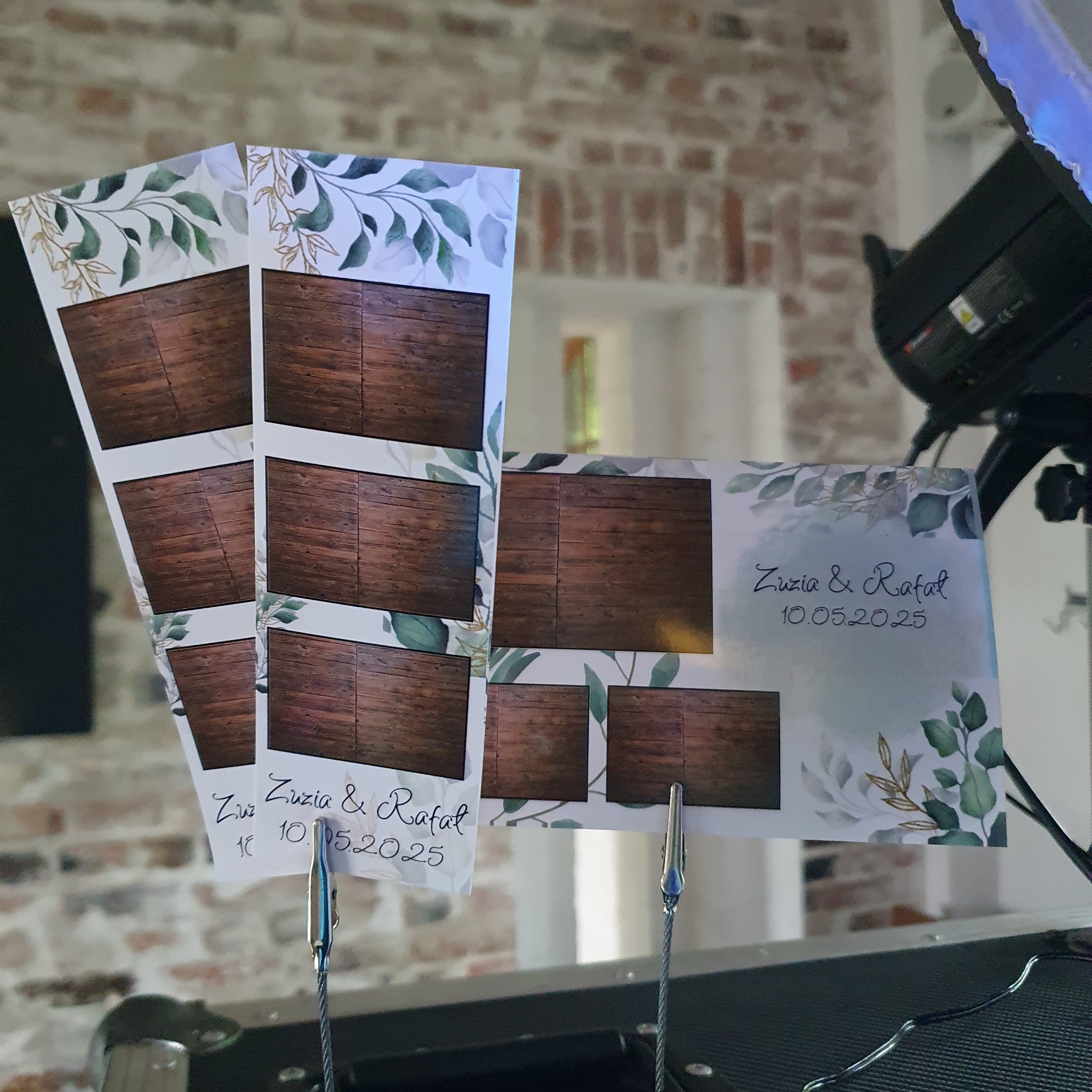 Paski zdjęć z fotobudki z motywem liści i datą ślubu, przypięte metalowymi klipsami. W tle widoczna ściana z cegły i sprzęt fotobudki.