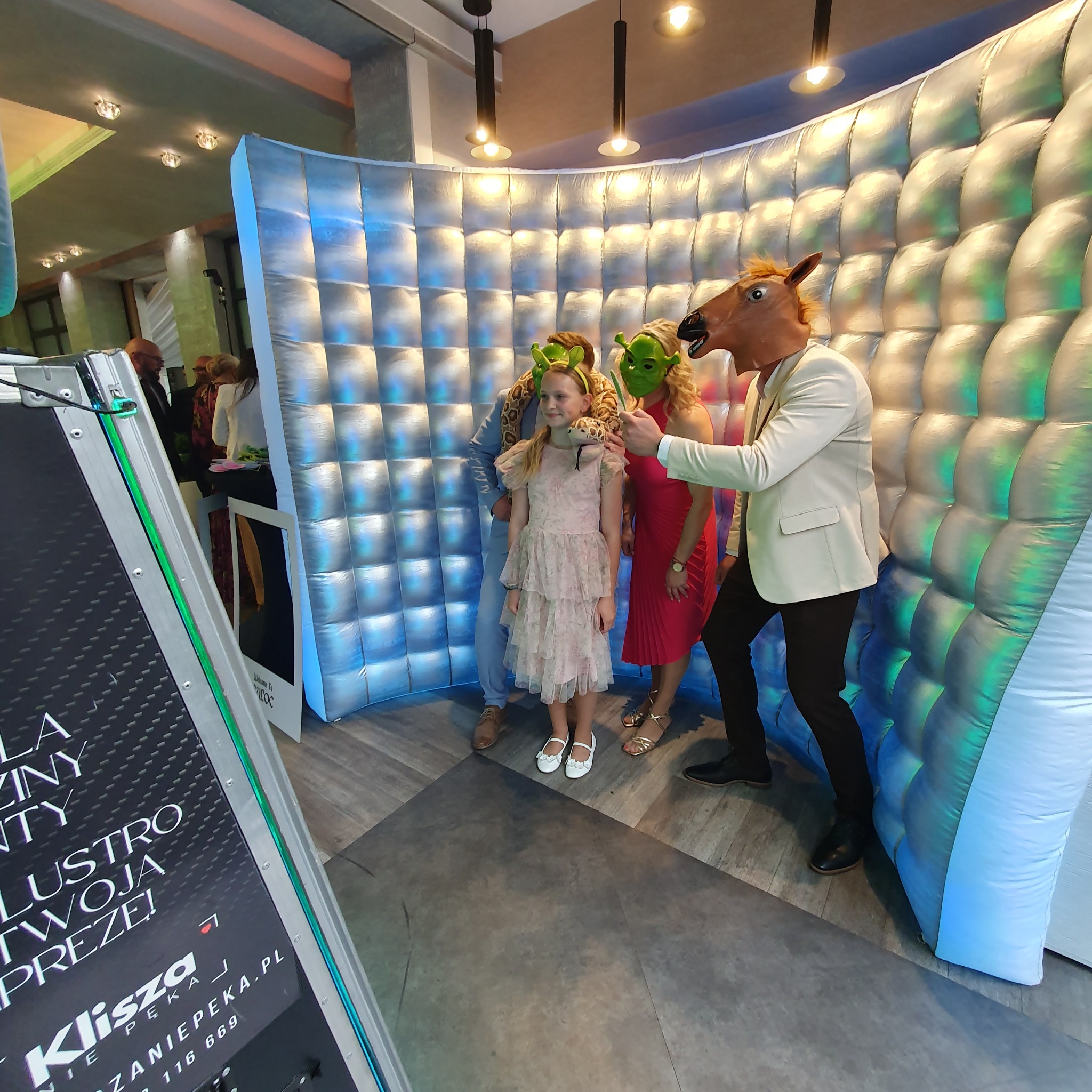 Grupa osób w maskach pozuje w dmuchanej fotobudce z efektem pikseli. Dziecko w sukience z zabawką węża. Widoczny fragment fotobudki z napisem. Nietypowe rekwizyty.