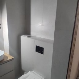 Sebastian Gawron - Nowoczesna łazienka w trakcie wykańczania: zabudowa stelaża WC z płytkami, widoczny przycisk spłukujący. Toaleta zabezpieczona folią.