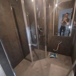 Sebastian Gawron - Nowoczesna kabina prysznicowa z ciemnymi płytkami, widoczne przyłącza wody i odbicie osoby w lustrze. Minimalistyczny design i szklane elementy.