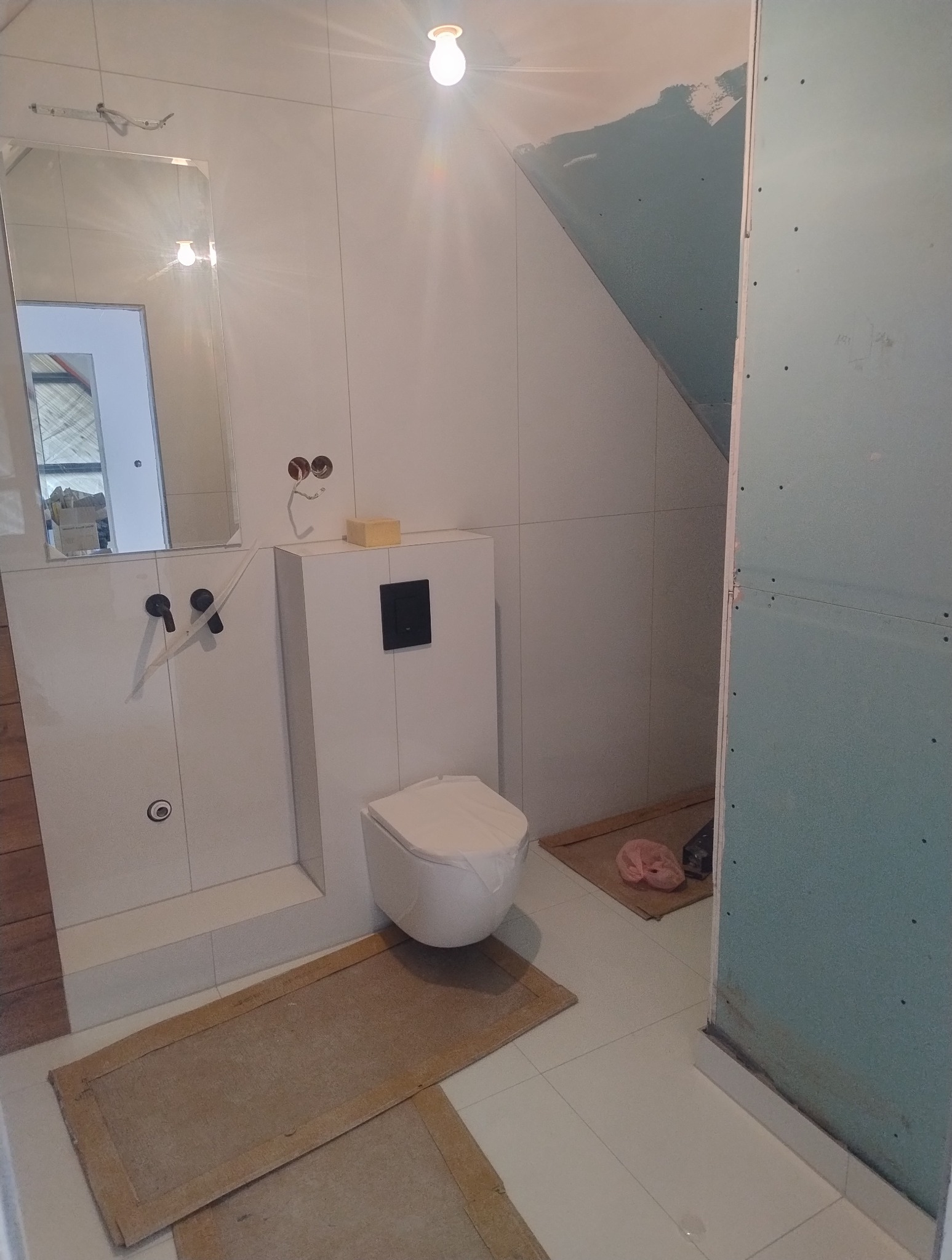 Biała łazienka w trakcie wykończenia: podwieszana toaleta, lustro, płytki ścienne, przygotowane ściany pod malowanie. Widoczne niedokończone detale.