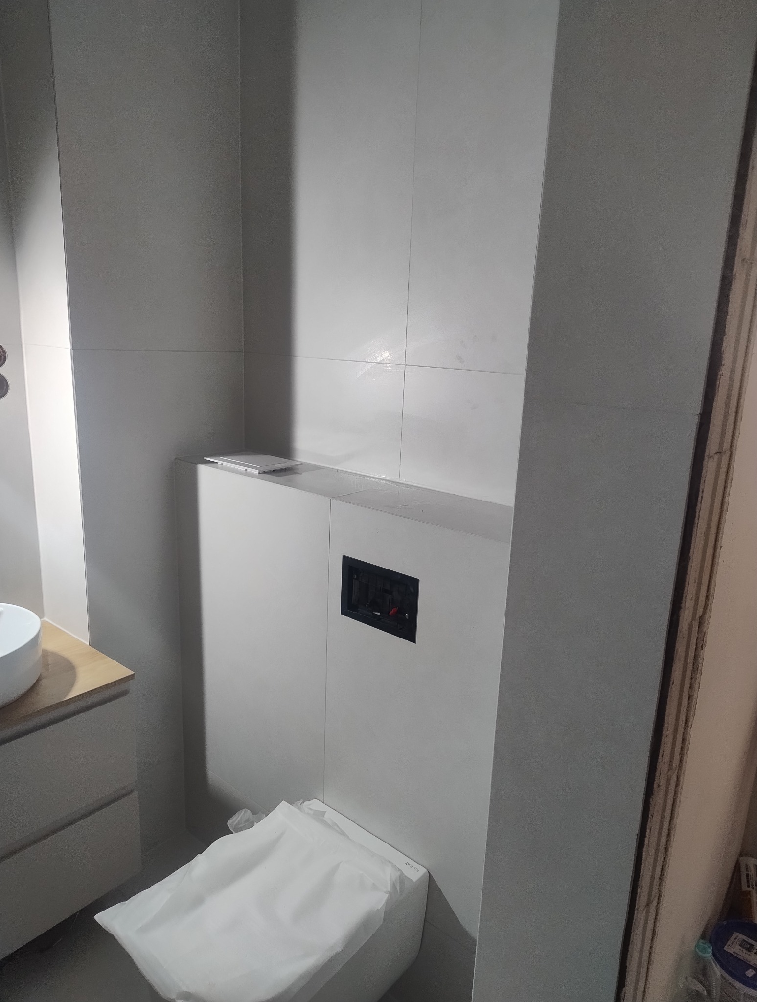 Nowoczesna łazienka w trakcie wykańczania: zabudowa stelaża WC z płytkami, widoczny przycisk spłukujący. Toaleta zabezpieczona folią.