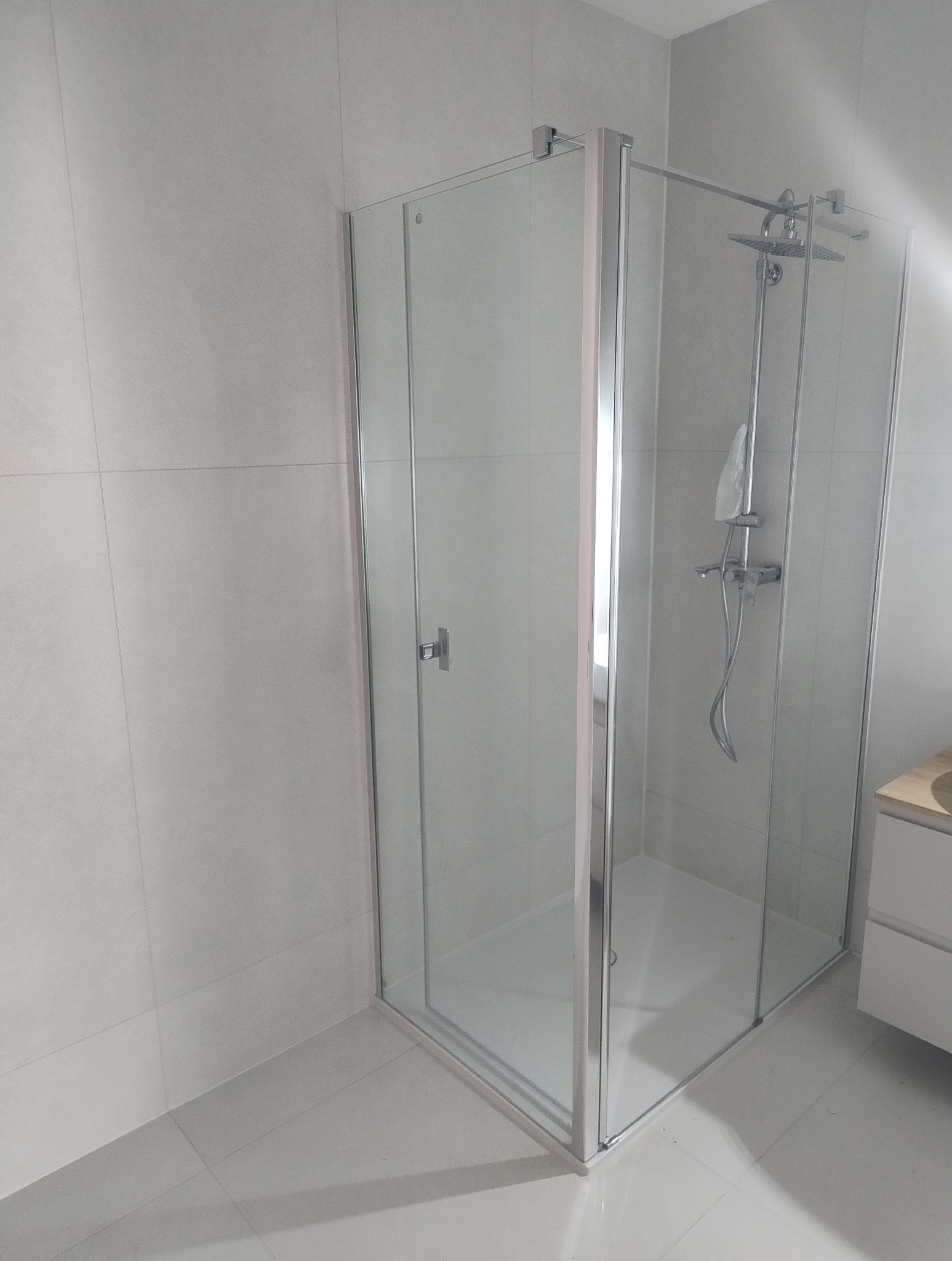 Nowoczesna kabina prysznicowa z chromowanymi profilami i deszczownicą, jasne płytki na ścianach i podłodze, minimalistyczny design.