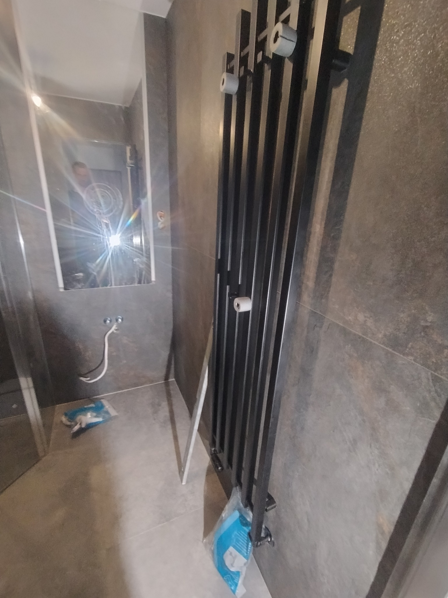 Nowoczesna łazienka z grzejnikiem drabinkowym, szarą glazurą i lustrem z odbiciem osoby. Widoczne przyłącza wodne i akcesoria montażowe. Wnętrze w trakcie wykańczania.