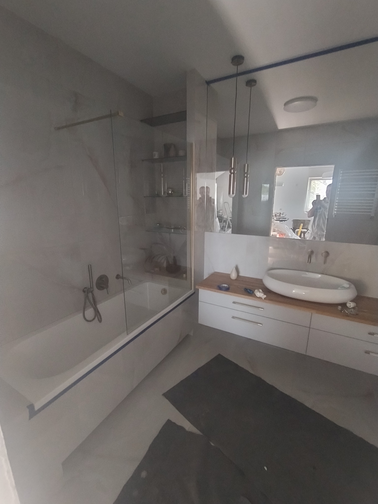 Nowoczesna łazienka z wanną za szklaną ścianką, umywalką na blacie i wiszącymi lampami. W tle widoczne półki i odbicie osoby robiącej zdjęcie.