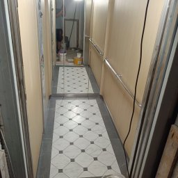 Drewniak Glazurnictwo - Wykończone wnętrze windy z nową podłogą w biało-szarą szachownicę i beżowymi panelami ściennymi. Widoczne metalowe poręcze i odbicie osoby w lustrze.