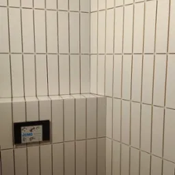 Białe, wąskie płytki ułożone pionowo w rogu łazienki. Widoczna instalacja podtynkowa WC i fragment ściany z półką. Minimalistyczne wykończenie.