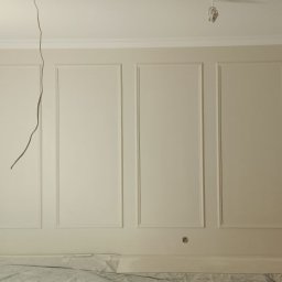 Drewniak Glazurnictwo - Ściana z dekoracyjnymi panelami i listwami w odcieniach bieli, zabezpieczone okno. W tle widoczna lampa oświetleniowa, kabel zwisający z sufitu.