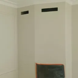 Wnętrze po malowaniu: kremowe ściany z dekoracyjną sztukaterią, zabezpieczony kominek i otwory wentylacyjne. Podłoga zakryta folią ochronną. Widok na wykończony róg pomieszczenia.