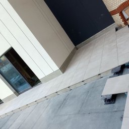 Drewniak Glazurnictwo - Jasne płytki tarasowe układane na regulowanych wspornikach, widok z góry na częściowo ukończoną powierzchnię przy budynku z oknami. Kartony w tle.