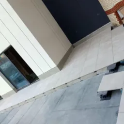 Jasne płytki tarasowe układane na regulowanych wspornikach, widok z góry na częściowo ukończoną powierzchnię przy budynku z oknami. Kartony w tle.