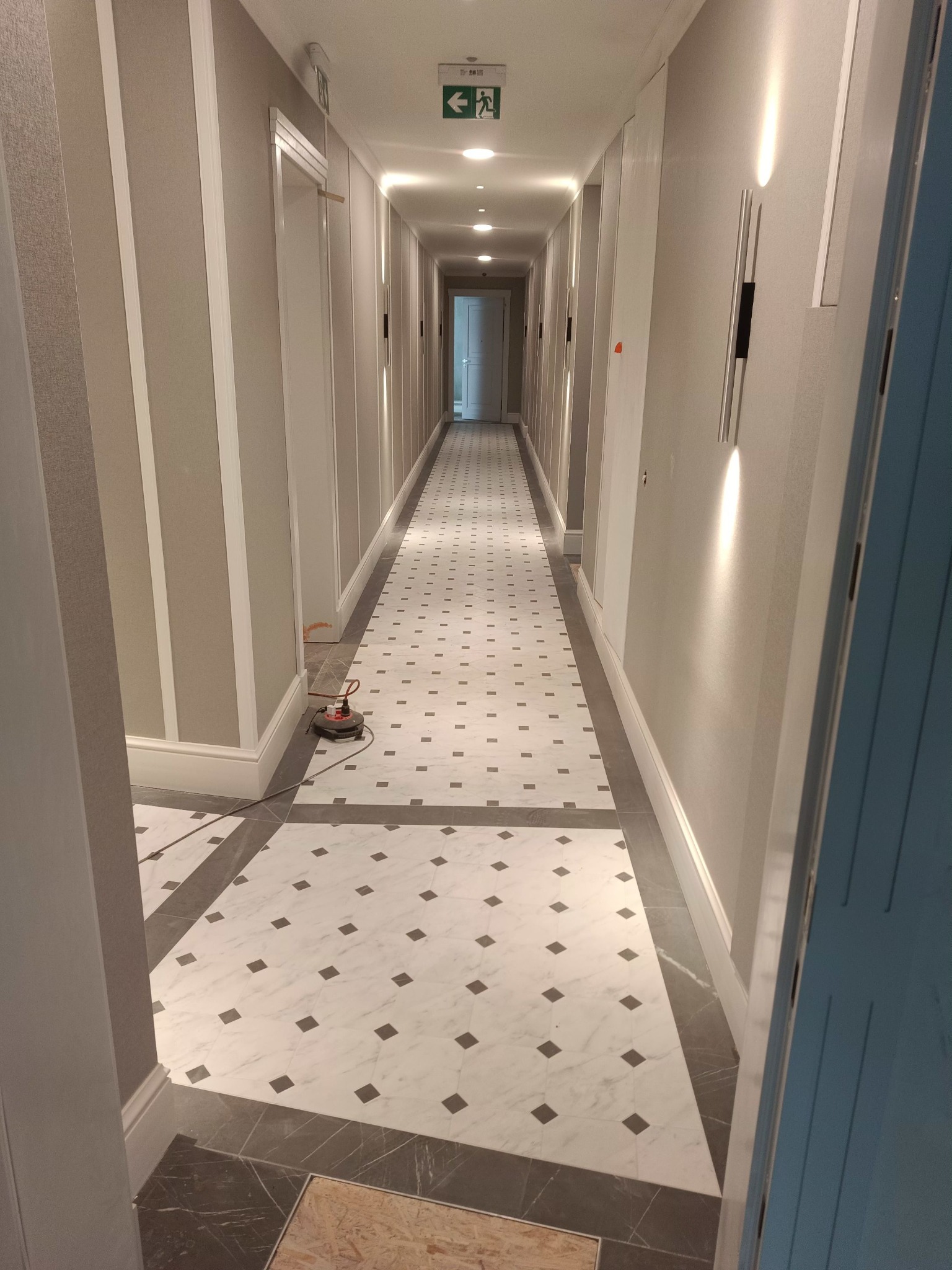 Długi korytarz z marmurową podłogą w biało-szare wzory, oświetlony lampami ściennymi. Widoczny sprzęt do czyszczenia podłóg. Wykończenie ścian w pionowe pasy.