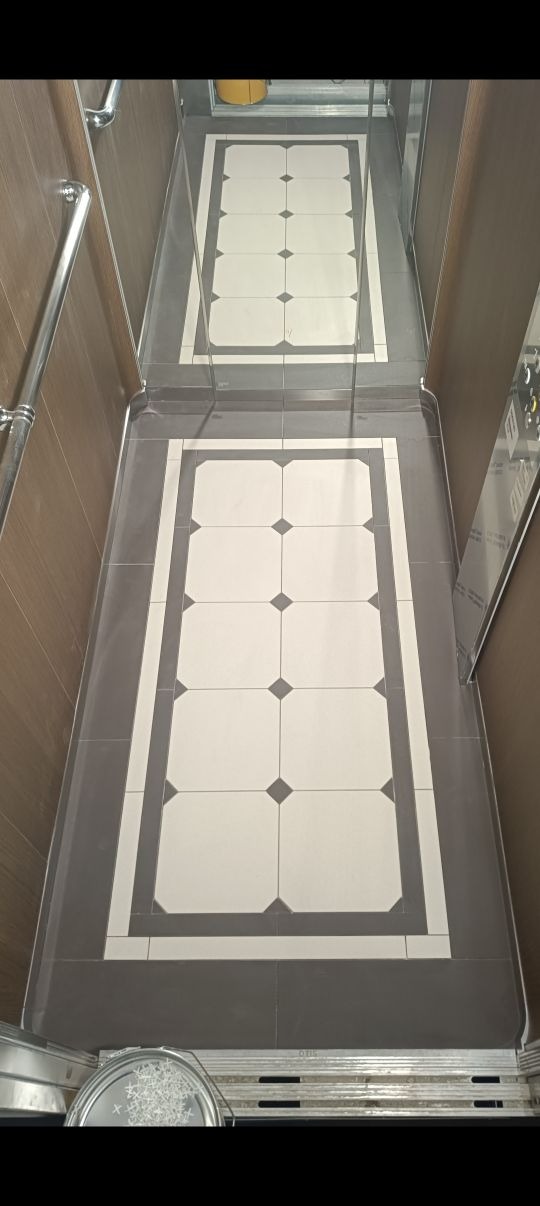 Wykończona podłoga w windzie z szarych i białych płytek w geometryczny wzór. Metalowe wykończenia i odblask w lustrze dodają elegancji.