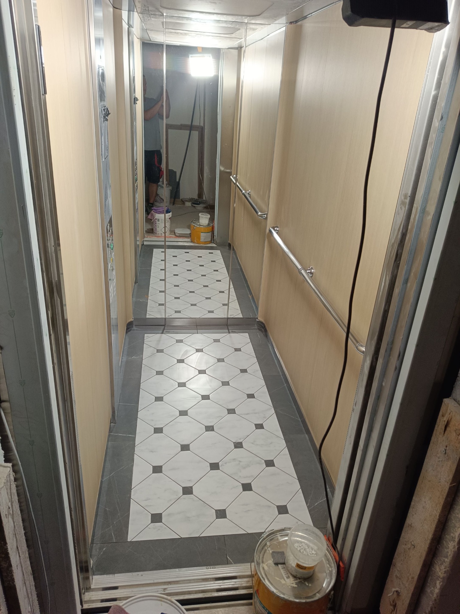 Wykończone wnętrze windy z nową podłogą w biało-szarą szachownicę i beżowymi panelami ściennymi. Widoczne metalowe poręcze i odbicie osoby w lustrze.