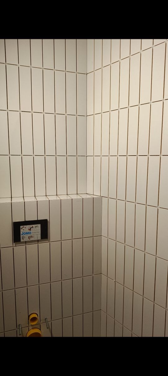 Białe, wąskie płytki ułożone pionowo w rogu łazienki. Widoczna instalacja podtynkowa WC i fragment ściany z półką. Minimalistyczne wykończenie.