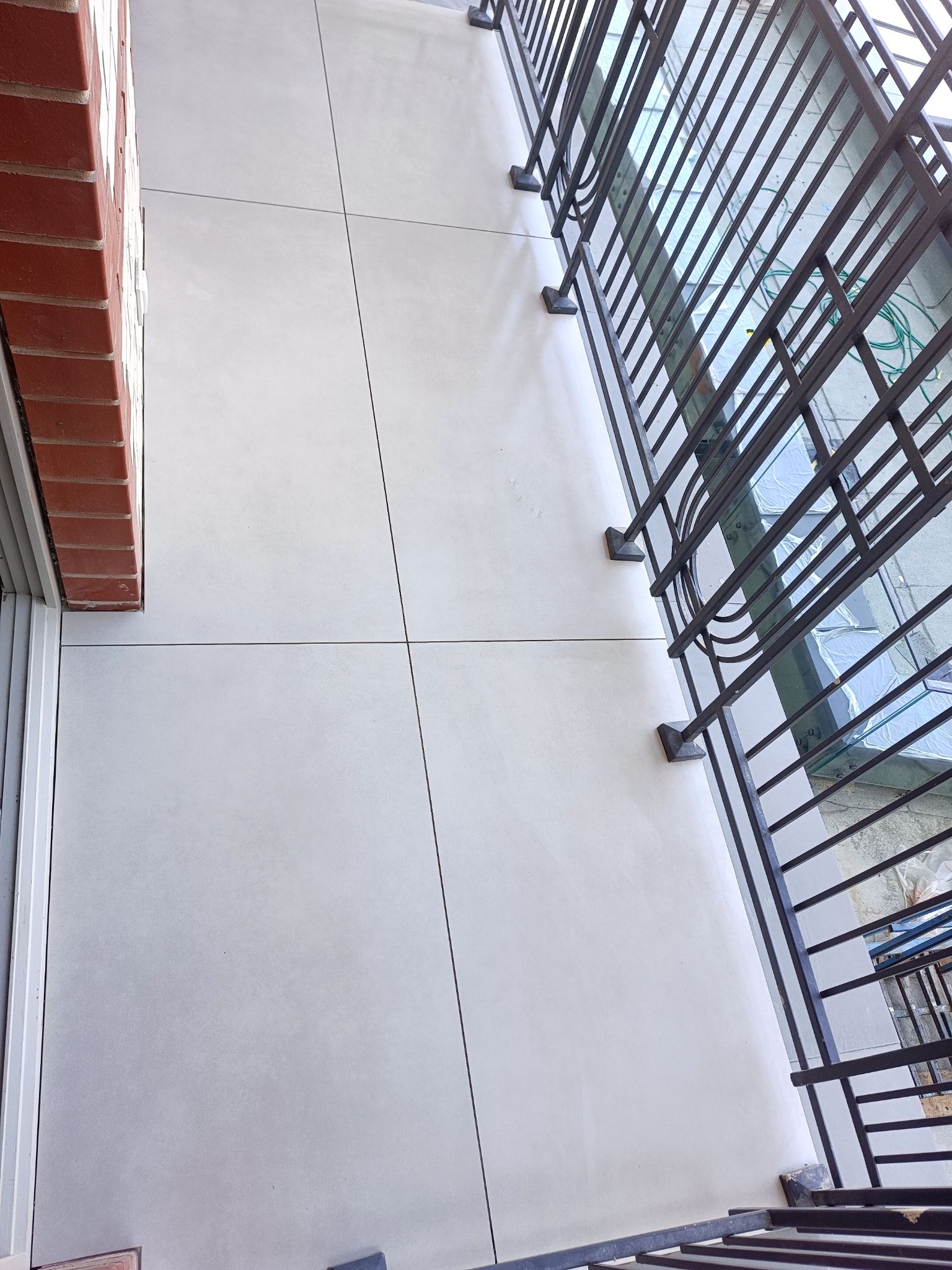 Jasnoszara, gładka posadzka balkonu wyłożona płytami, widok z góry, z fragmentem ceglanej ściany i metalowej balustrady. Minimalistyczny design, świeżo po remoncie.