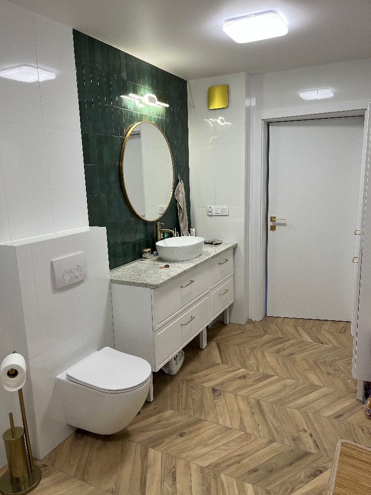 Nowoczesna łazienka z zielonymi płytkami na ścianie, owalnym lustrem w złotej ramie i drewnianą podłogą ułożoną w jodełkę. Biała szafka pod umywalką i podwieszana toaleta.