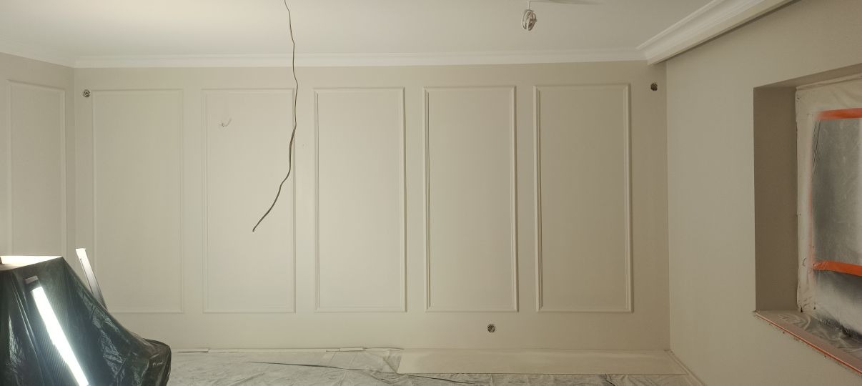 Ściana z dekoracyjnymi panelami i listwami w odcieniach bieli, zabezpieczone okno. W tle widoczna lampa oświetleniowa, kabel zwisający z sufitu.