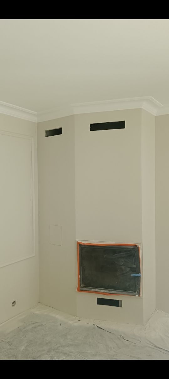 Wnętrze po malowaniu: kremowe ściany z dekoracyjną sztukaterią, zabezpieczony kominek i otwory wentylacyjne. Podłoga zakryta folią ochronną. Widok na wykończony róg pomieszczenia.