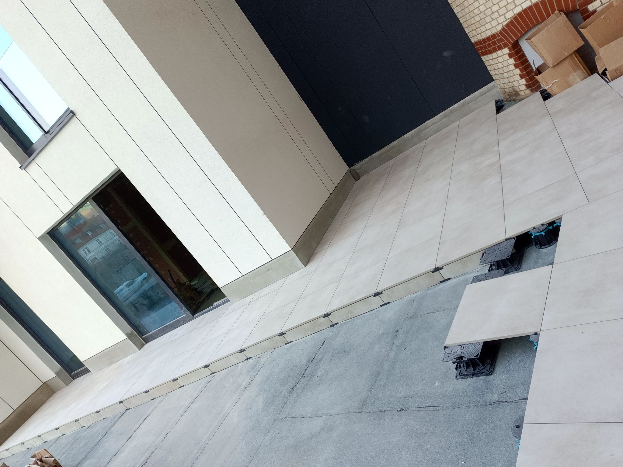 Jasne płytki tarasowe układane na regulowanych wspornikach, widok z góry na częściowo ukończoną powierzchnię przy budynku z oknami. Kartony w tle.