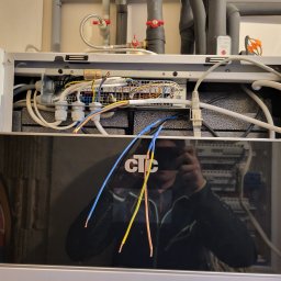 FHU Maciej Malinowski Intertrans - Otwarta obudowa kotła grzewczego z widocznymi przewodami elektrycznymi i elementami instalacji. Widoczny panel sterowania i odbicie osoby robiącej zdjęcie.