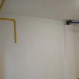 Biała ściana z żółtą instalacją gazową i niebieską instalacją wodną, widoczne gniazdka elektryczne i fragment podłogi. Świeżo po szpachlowaniu.