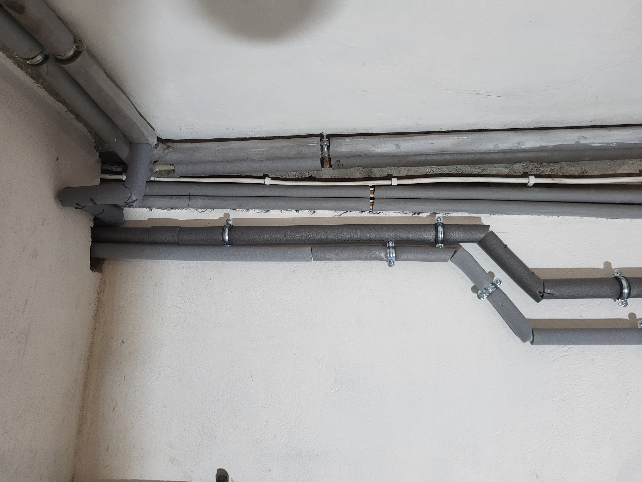 Szare, zaizolowane rury poprowadzone wzdłuż sufitu i ściany, zamocowane metalowymi uchwytami. Izolacja miejscami niedokładna. Widoczne kable elektryczne.