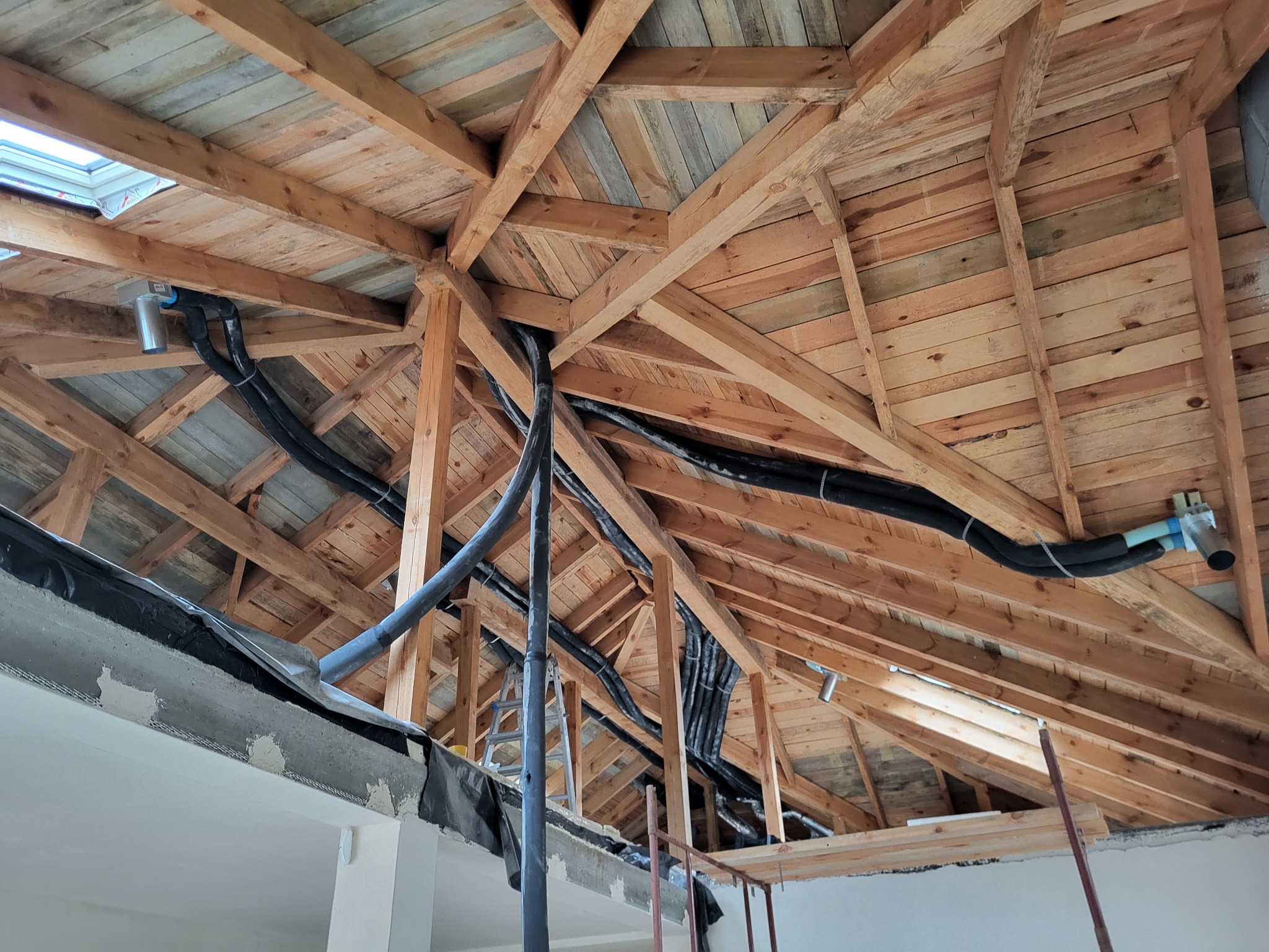 Drewniana konstrukcja dachu z widocznymi elementami instalacji wentylacyjnej i oknem dachowym. Fragment ściany w trakcie prac wykończeniowych.