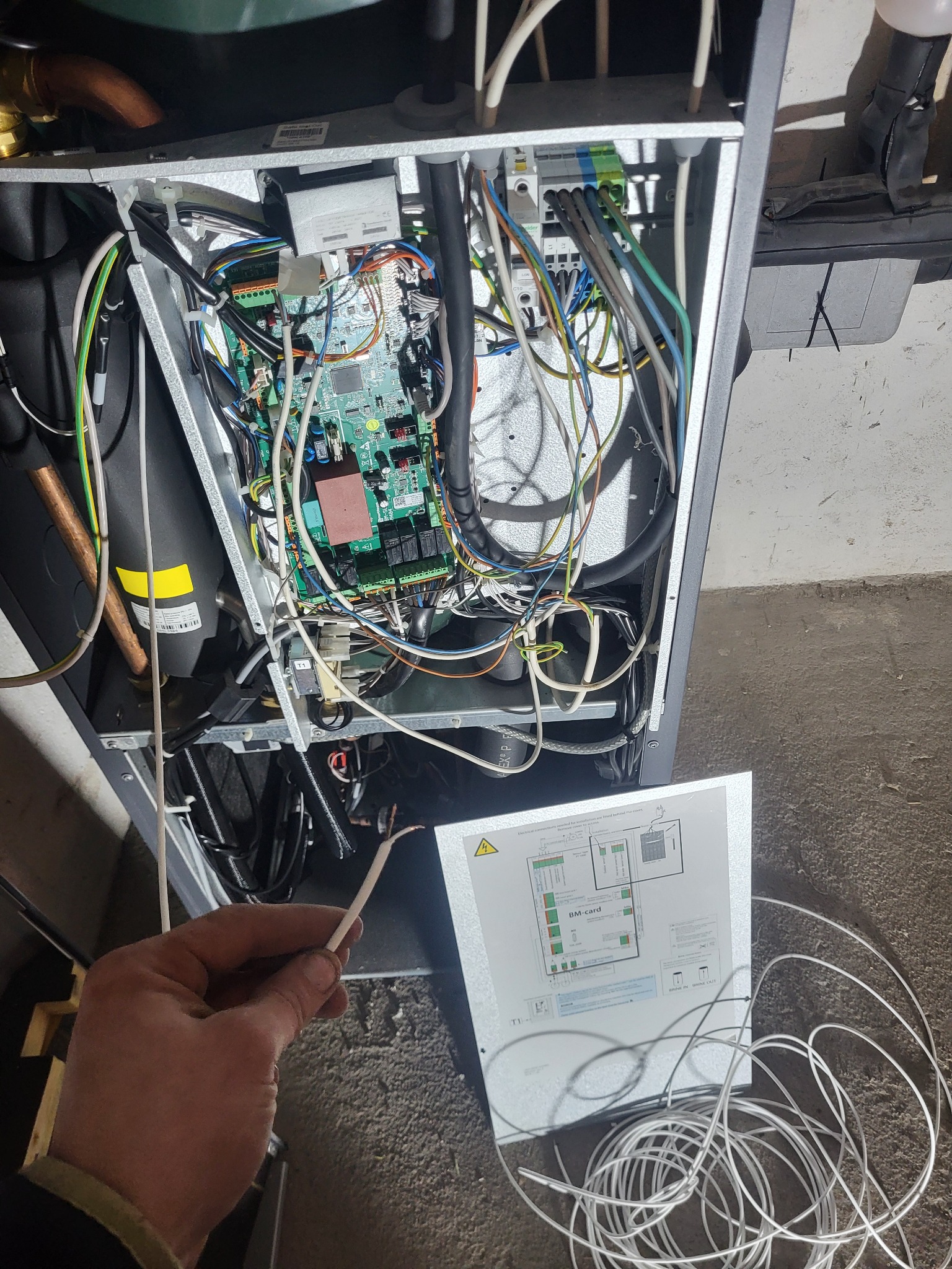 Otwarta obudowa urządzenia z widoczną płytą główną i plątaniną kabli. W dłoni trzymany przewód, obok schemat połączeń na metalowej obudowie.