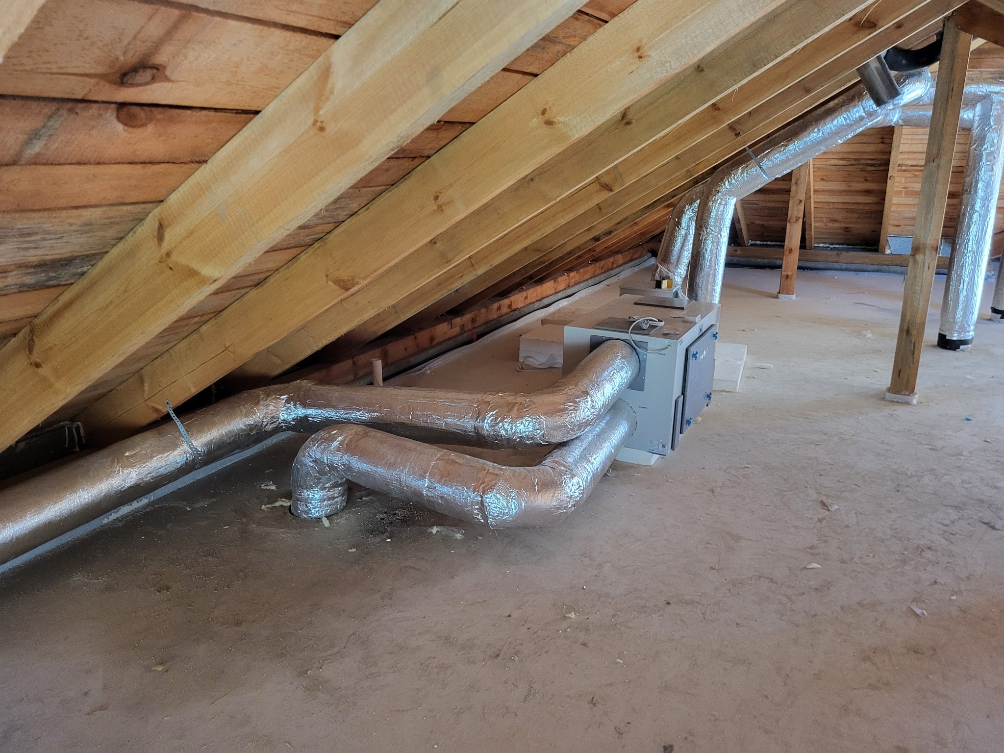 Szare, izolowane rury wentylacyjne pod skośnym, drewnianym dachem na poddaszu. Widoczny centralny moduł wentylacyjny i drewniane belki konstrukcyjne. Jasne oświetlenie.