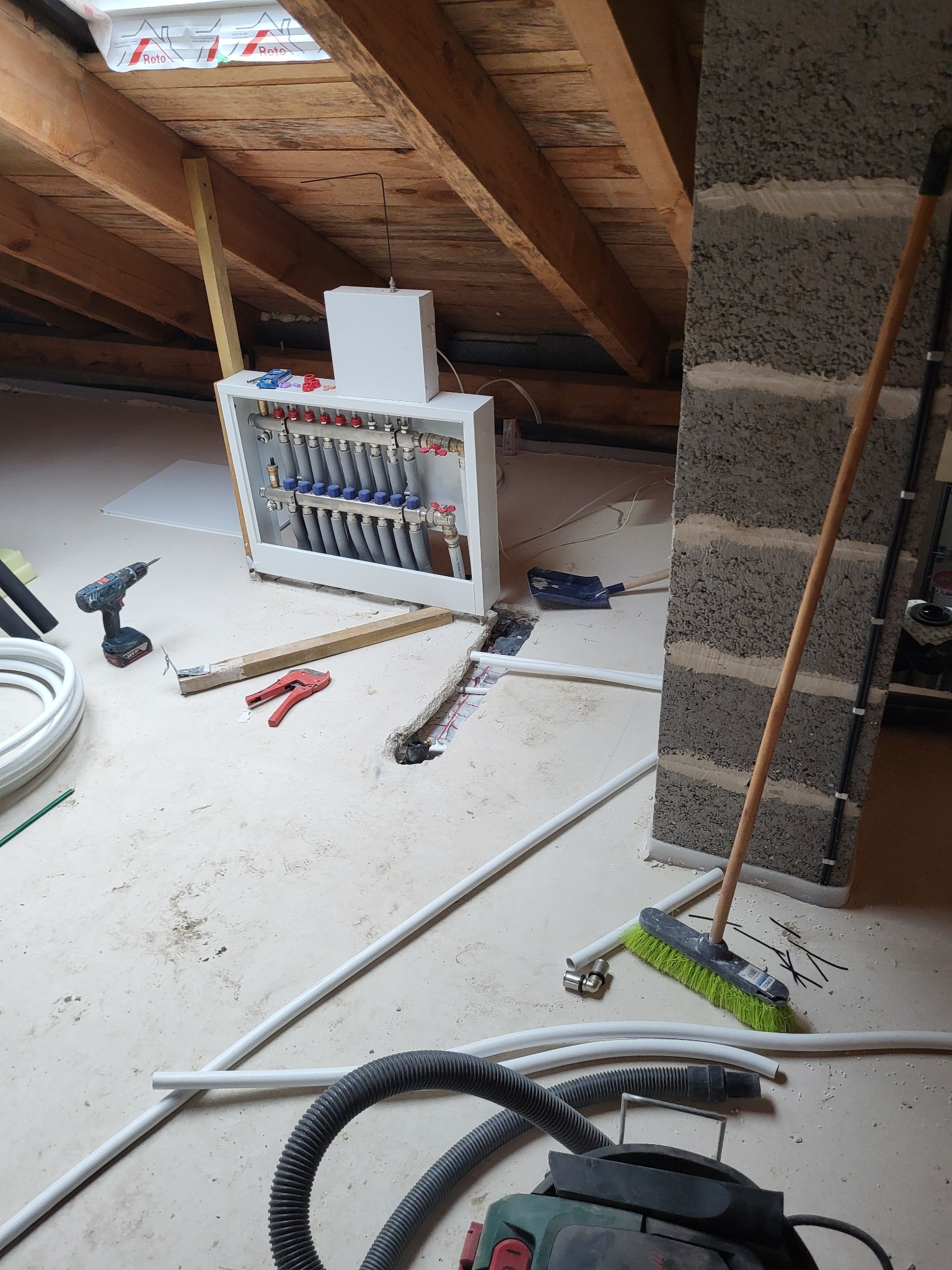 Rozdzielacz ogrzewania podłogowego w trakcie montażu na poddaszu. Widoczne rury, narzędzia i elementy instalacji. Oświetlenie naturalne z okna dachowego.