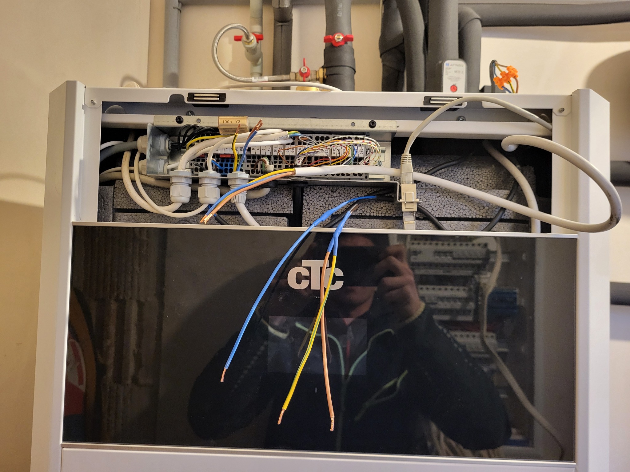 Otwarta obudowa kotła grzewczego z widocznymi przewodami elektrycznymi i elementami instalacji. Widoczny panel sterowania i odbicie osoby robiącej zdjęcie.