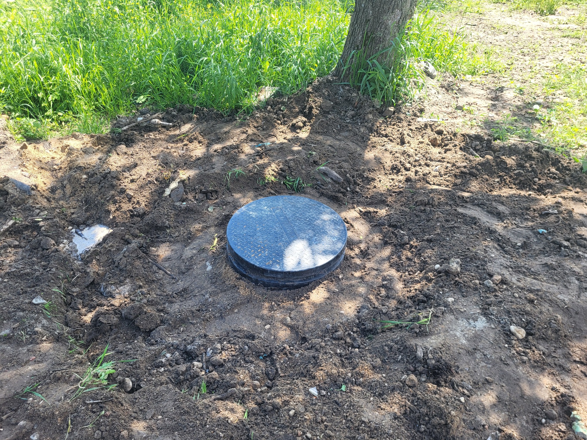 Okrągła, czarna pokrywa włazu kanalizacyjnego osadzona w ziemi, obok pnia drzewa i trawy. Prace ziemne w toku, przygotowanie terenu pod instalację.